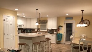 A kitchen with white cabinets, a central island with stools, a dining table with chairs, and pendant lighting. - I LOVE BEACH MUSIC (Ocean Isle Beach)