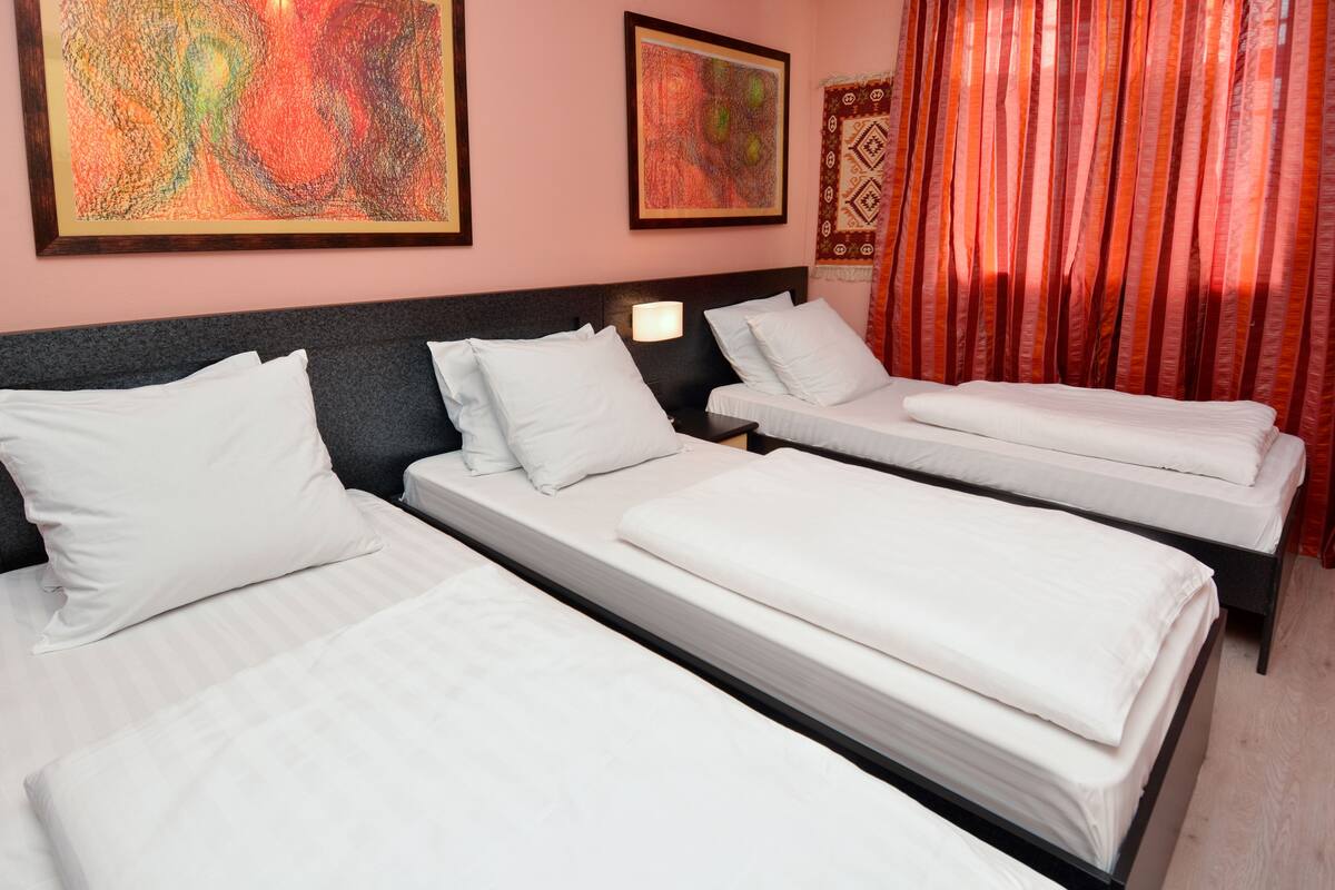 Habitación de hotel con tres camas, cada una con ropa de cama y almohadas blancas. Hay dos lámparas de pared en las mesitas de noche.