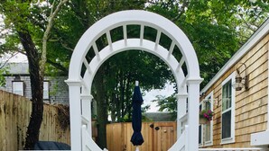 Un portón blanco de madera con arco semicircular, flanqueado por macetas blancas con flores, ubicado en un camino de grava.