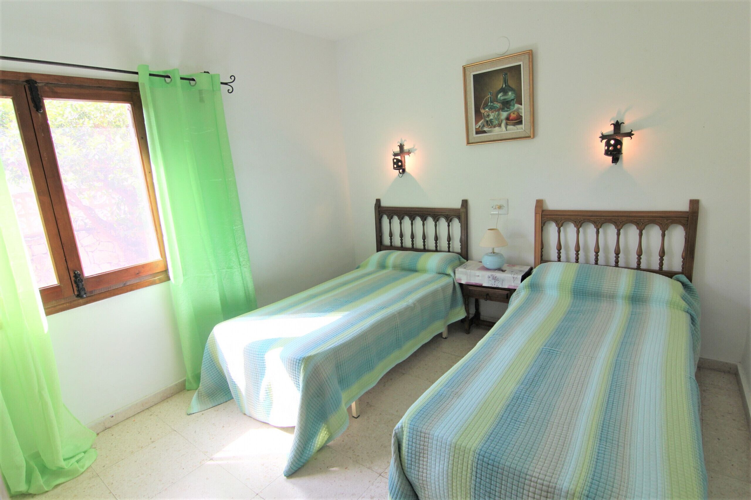 Una habitación con dos camas individuales, una ventana con cortinas, un cuadro en la pared y dos luces fijadas a la pared.