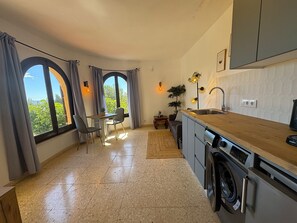 Cocina compacta con lavadora integrada, comedor con mesa y sillas, y ventanas amplias con cortinas.