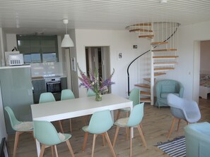 Une cuisine moderne dotée d’un four et d’un micro-ondes encastrés, un coin repas avec une table blanche et des chaises vert clair, et un escalier en colimaçon menant à un niveau supérieur.