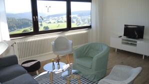 A modern living room with a sofa, two chairs, a glass coffee table, a television, and a view of a green landscape through large windows. - Renovated, bright, light-flooded 4-room apartment (Saig)