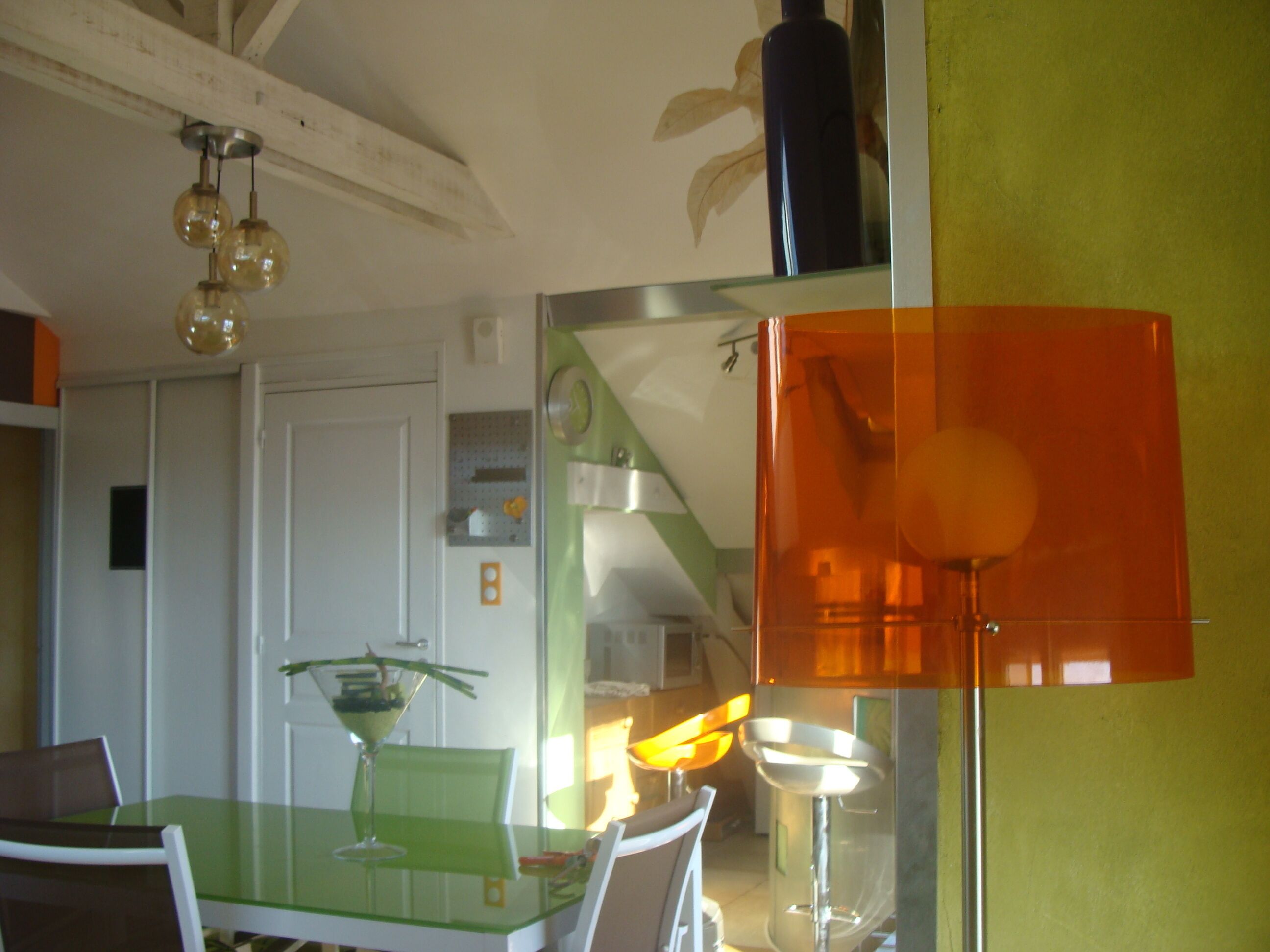 A dining area with a green table, chairs, and a glass-top table. There is a mirror on the wall, a ceiling light fixture, and a door in the background.