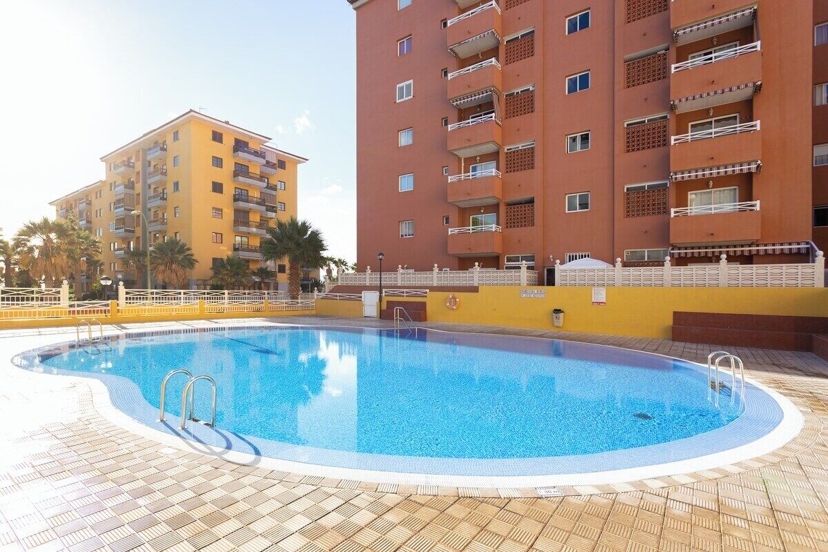 A swimming pool surrounded by a tiled deck, with a yellow building and a red building in the background.