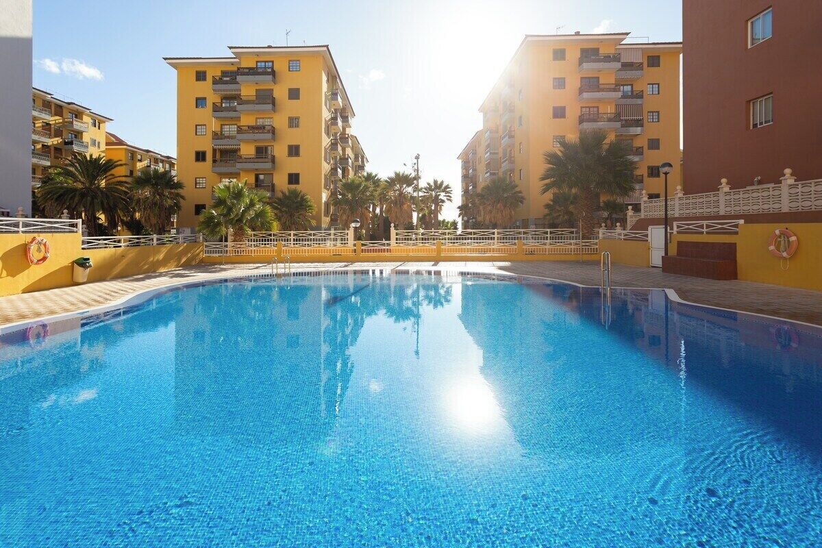 A swimming pool surrounded by yellow buildings with balconies, palm trees, and a lifebuoy on the wall.