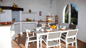 Un patio iluminado por el sol con una mesa de comedor lista para cuatro personas, rodeada de sillas blancas. Se observa un pequeño área de cocina con un refrigerador y una repisa con utensilios.