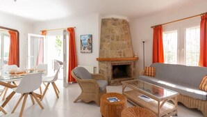 Una sala de estar con chimenea de piedra, una zona de comedor con sillas blancas y un sofá con cortinas naranjas.