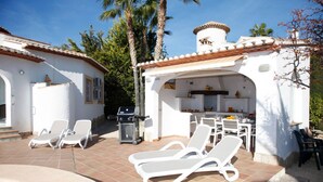 Un área de piscina con sillones de descanso, una parrilla y una cocina con mesa y sillas.