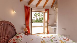 Un dormitorio con cama, una silla de mimbre, una mesita de noche, una lámpara, una ventana con cortinas y vistas a palmeras.