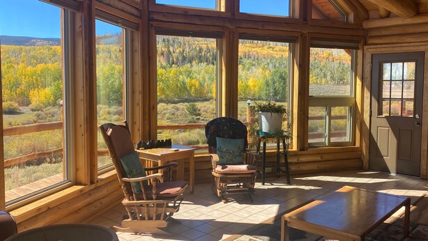 Une cabane en rondins dotée de grandes fenêtres qui offrent une vue sur le feuillage automnal. À l’intérieur, on trouve des chaises en bois, une table et une plante en pot.