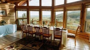 Une cabane en rondins dotée d’un coin repas avec une table dressée pour quatre personnes, entourée de grandes fenêtres offrant une vue sur la campagne.