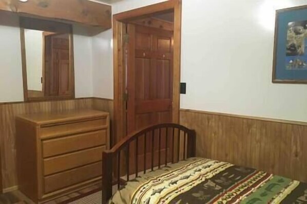 A bedroom with a wooden dresser, a bed with a patterned bedspread, a wooden chair, and a wooden door.