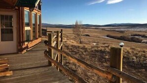 Une terrasse en bois mène à un bâtiment doté de grandes fenêtres, qui offre une vue sur un paysage plat avec des collines à l’horizon.