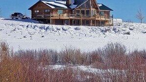 Une grande cabane en rondins, dotée de nombreuses fenêtres et d’un porche couvert, située sur une colline enneigée.