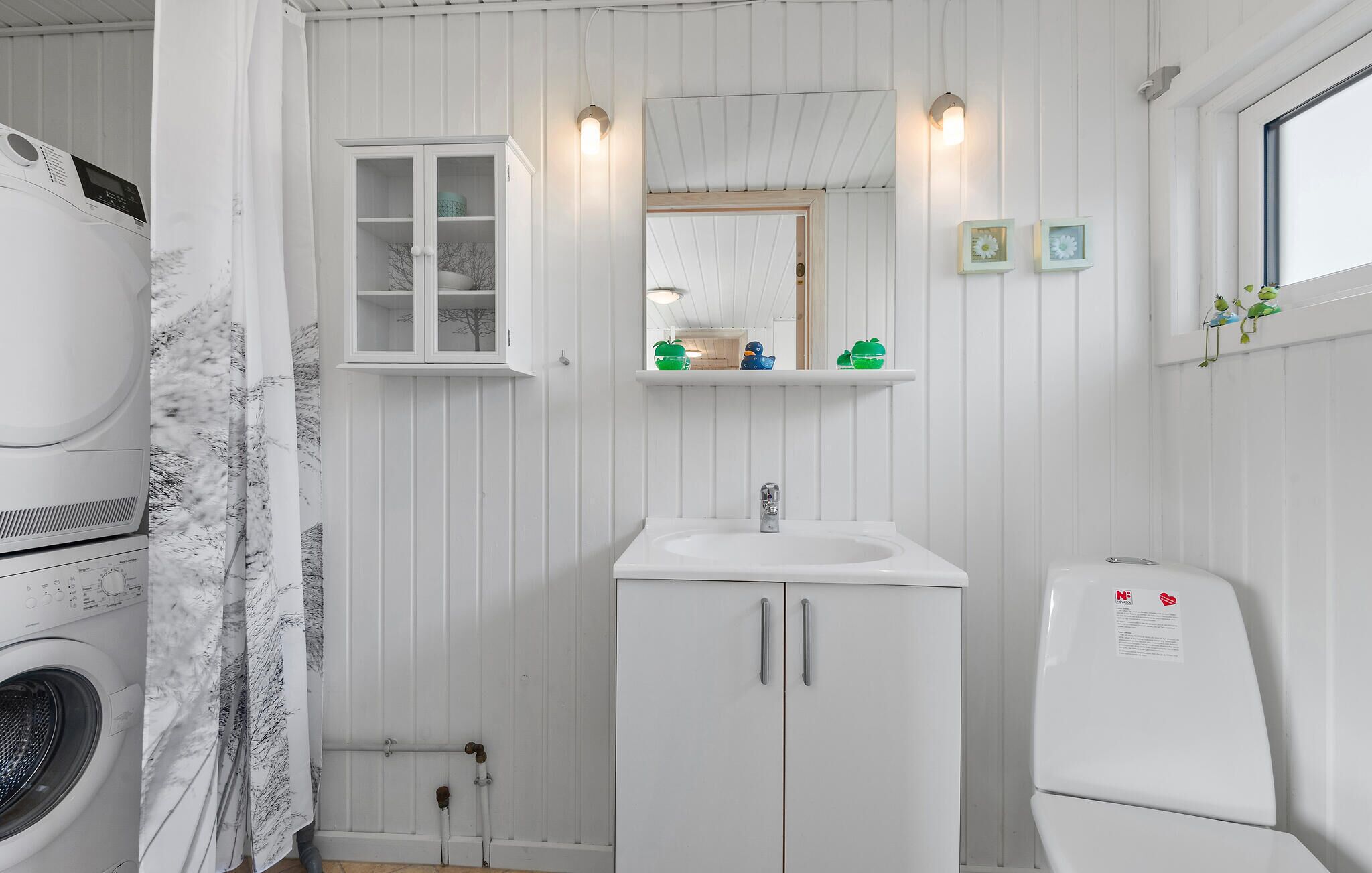 Une petite salle de bain équipée d’un lave-linge, d’un évier et de toilettes. On y trouve un meuble avec un miroir au-dessus de l’évier et une petite étagère où sont disposés des objets décoratifs.