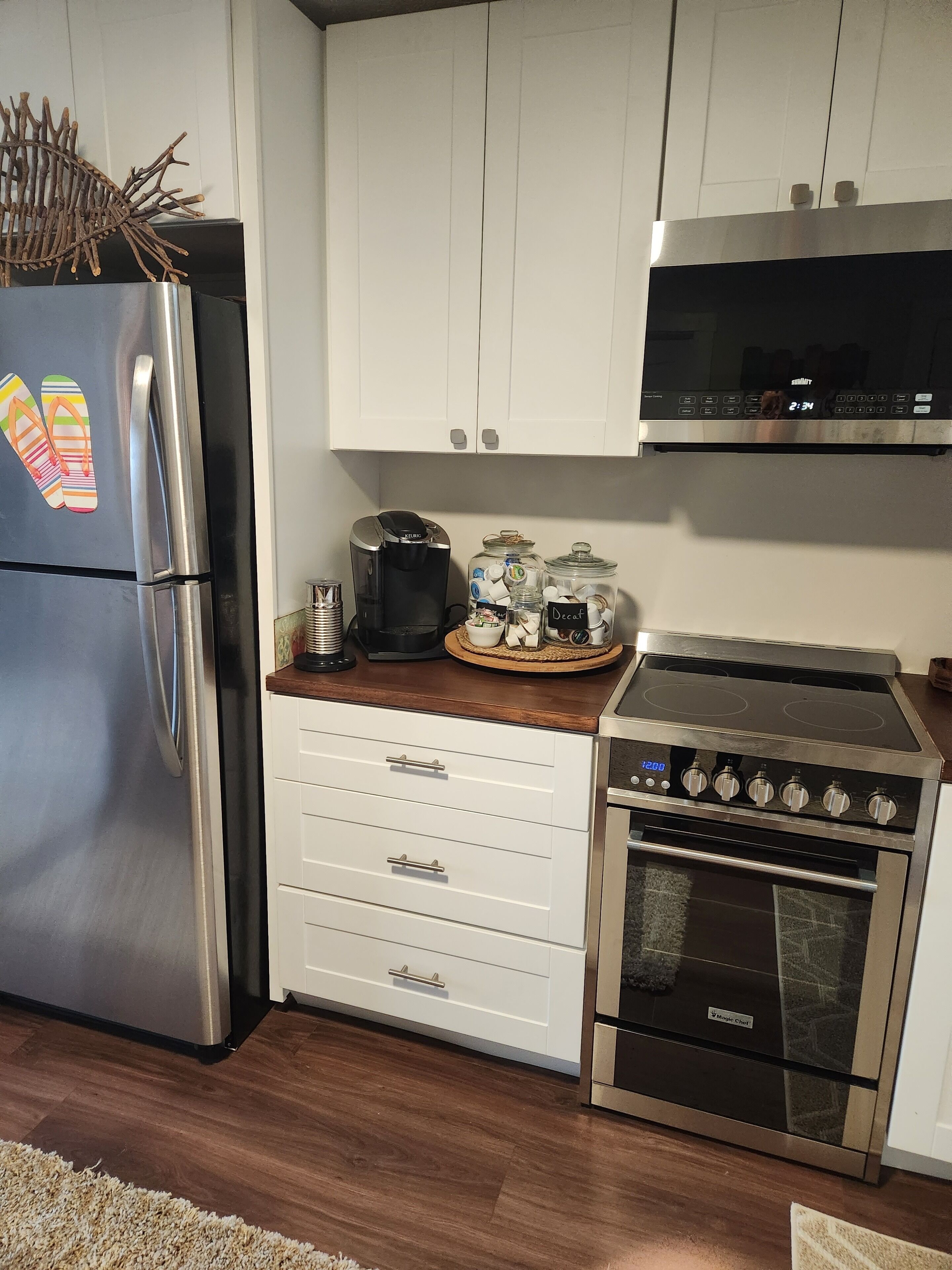Una cocina con un refrigerador de acero inoxidable, una cafetera, un microondas, una estufa y un juego de gabinetes blancos.