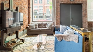 Un salon avec un mur en briques, un canapé, un téléviseur et une fenêtre donnant sur des immeubles.