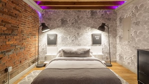 Une chambre avec un mur en briques, un lit avec une tête de lit blanche, deux tables de chevet avec des lampes et un mur recouvert d’un papier peint à motifs de nuages.
