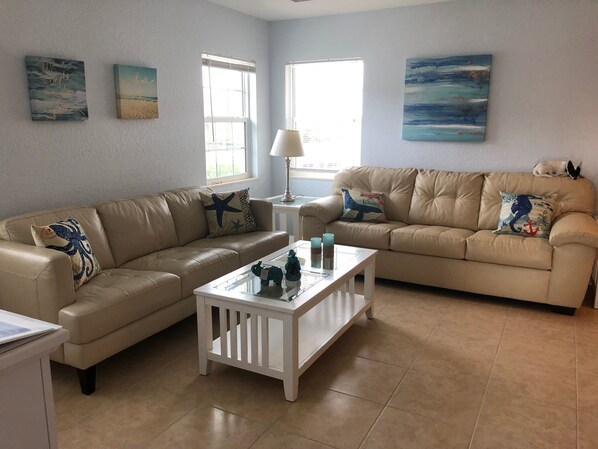 A living room with two beige sofas, a glass coffee table, and a lamp. There are wall art pieces and a window with blinds.