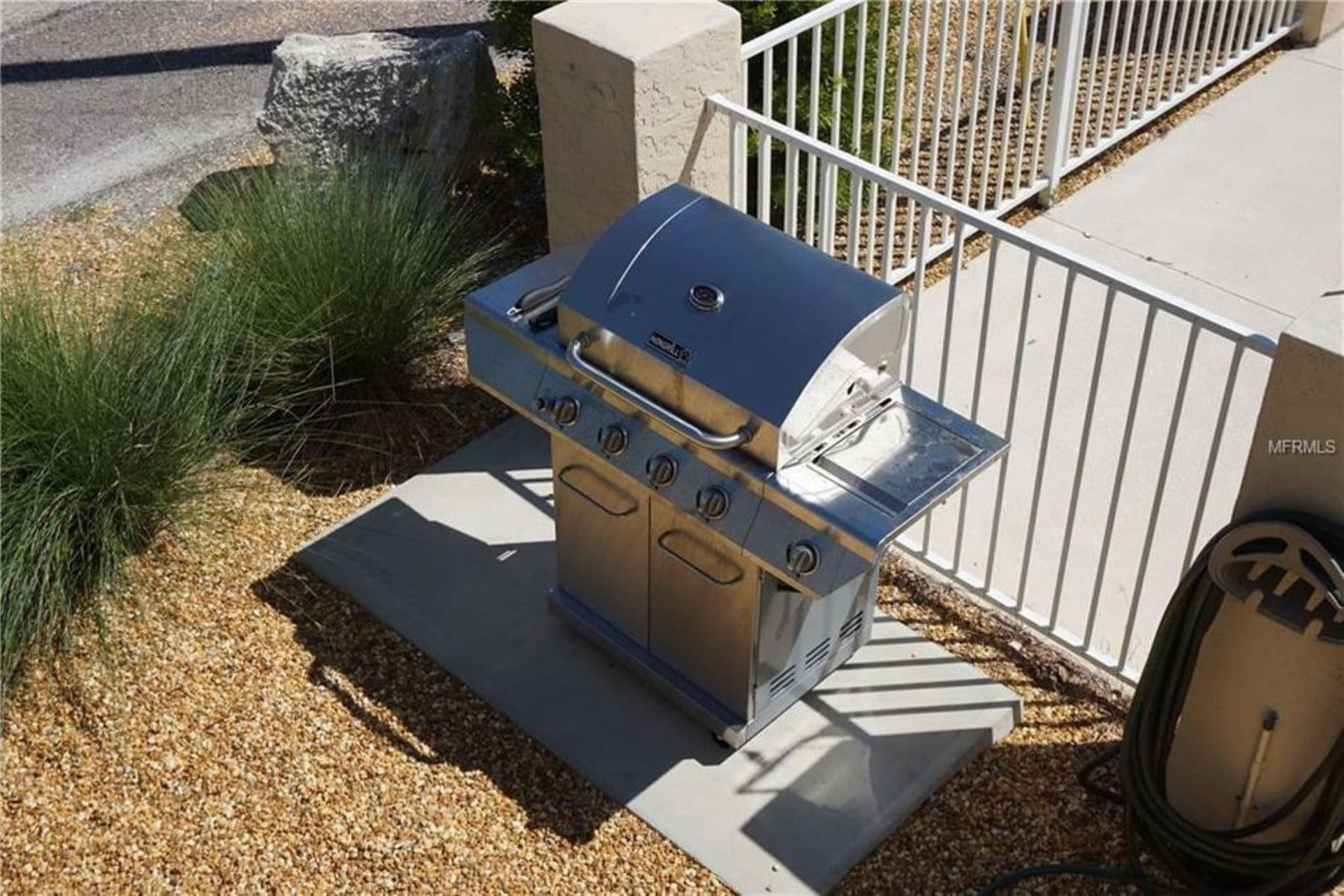 Una parrilla de acero inoxidable portátil con tapa y cuatro quemadores, ubicada sobre una base de concreto al lado de una cerca de metal blanco.