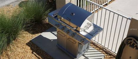 Una parrilla de acero inoxidable portátil con tapa y cuatro quemadores, ubicada sobre una base de concreto al lado de una cerca de metal blanco.