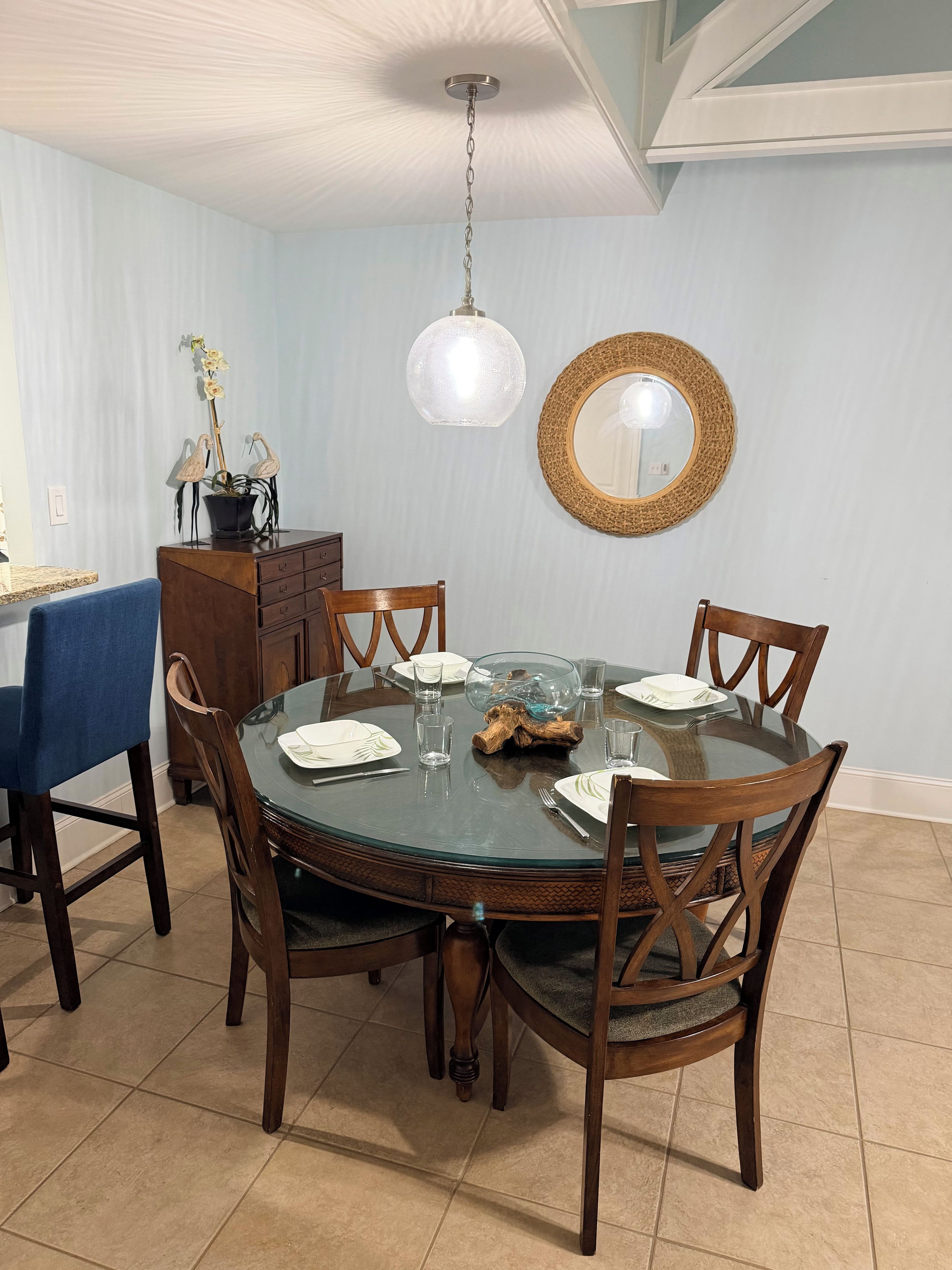 Een eetruimte met een ronde glazen tafel, houten stoelen, een blauwe tafelkleed, een houten kast, een hanglamp en een geweven spiegel aan de muur.