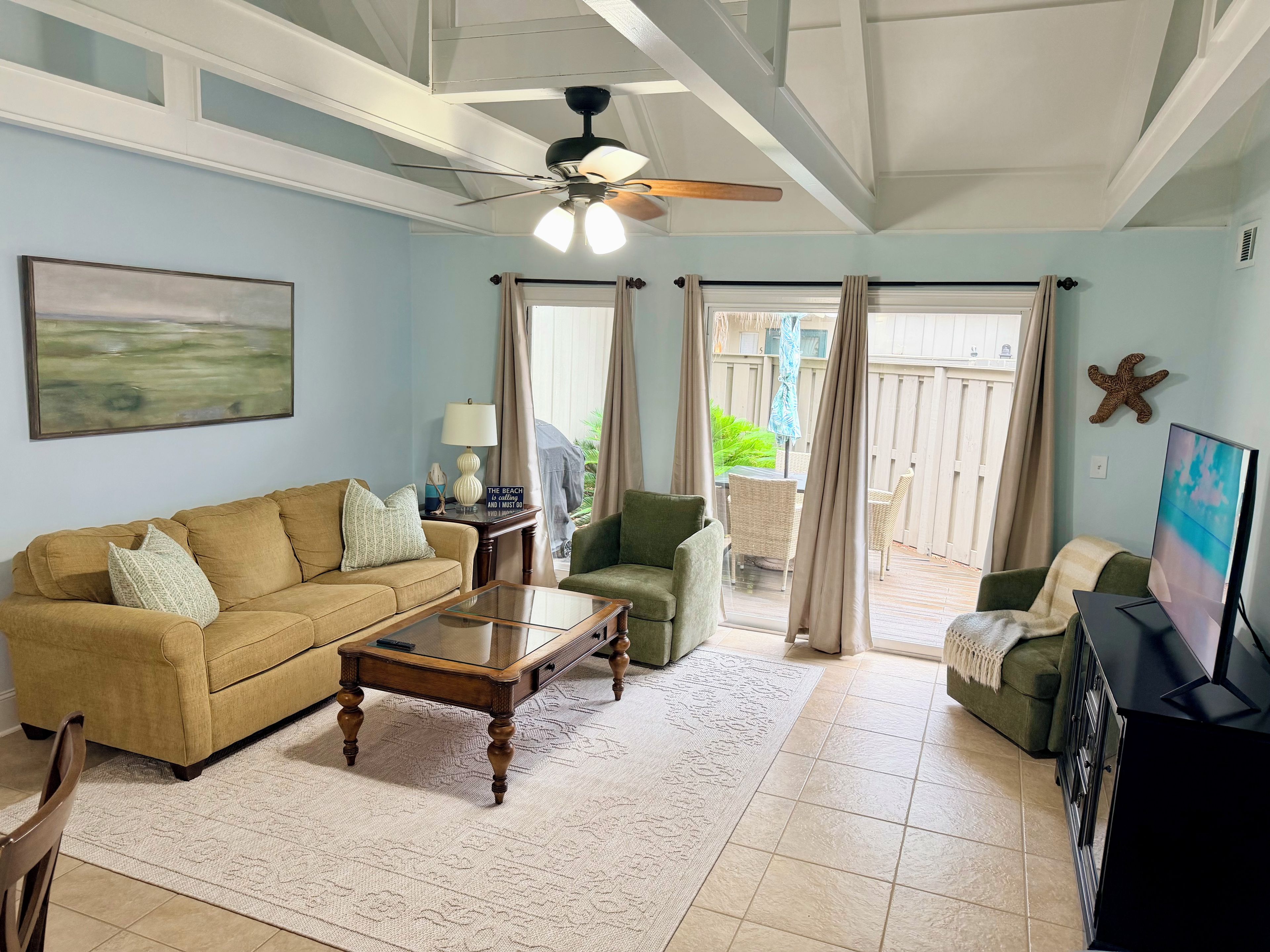 A living room with a beige sofa, a green armchair, a coffee table, a TV, and a ceiling fan.