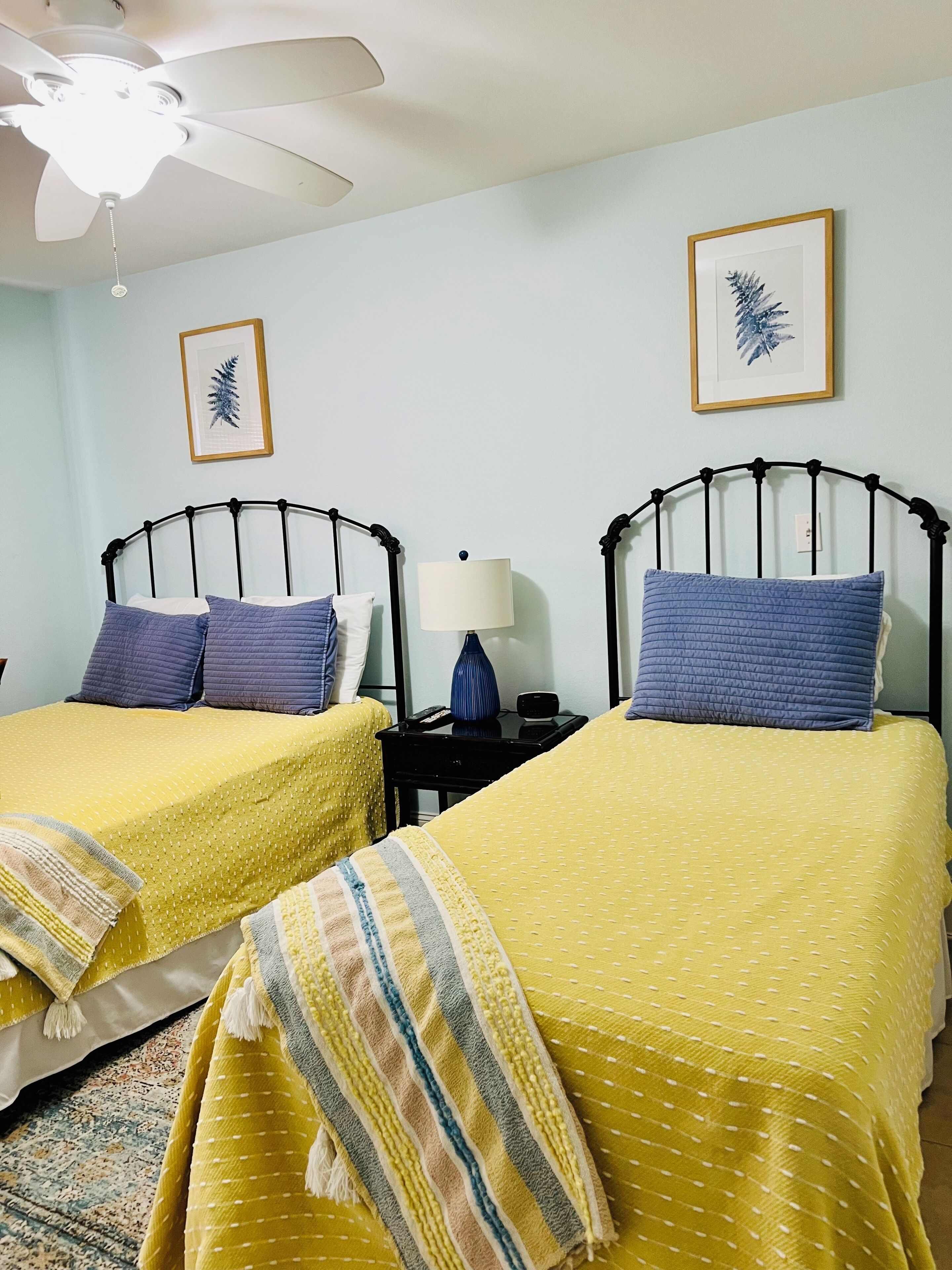 Dos camas individuales con ropa de cama amarilla, un ventilador de techo, una mesita de noche con lámpara y cuadros en la pared.