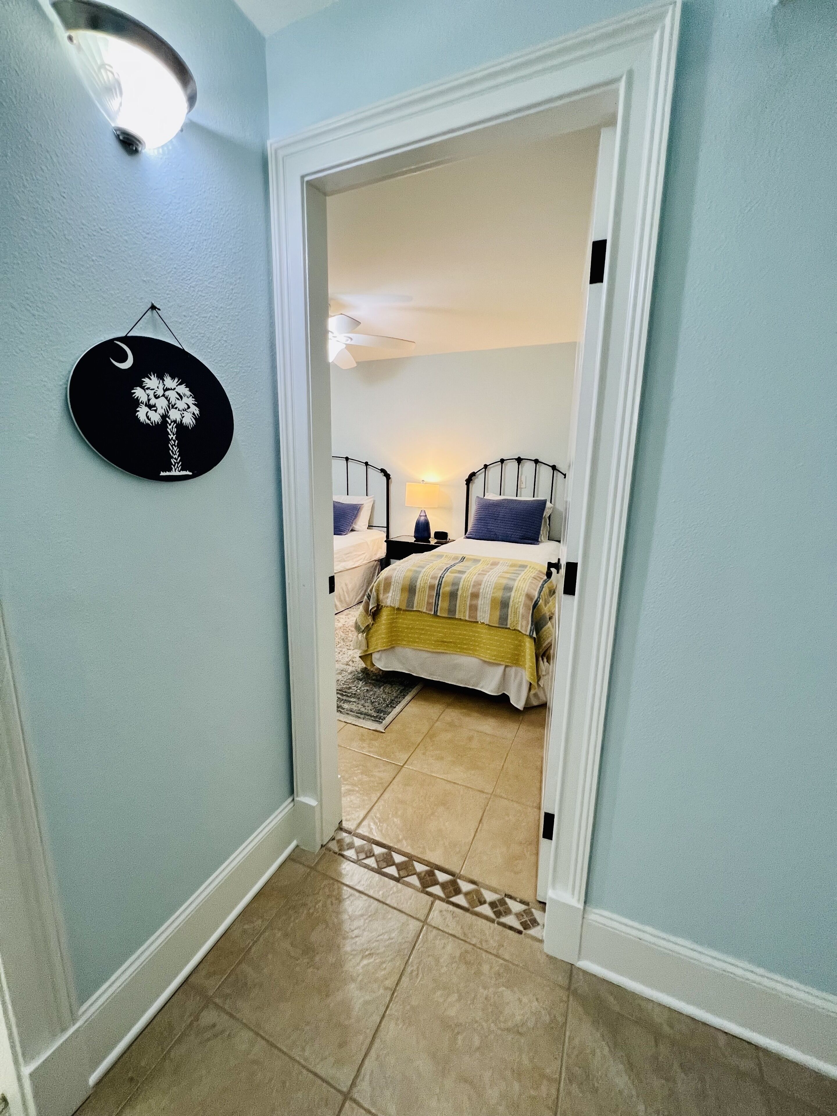 A hallway leading to a bedroom with two beds, a ceiling fan, and a wall-mounted light fixture.