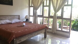 A bedroom with a wooden bed, white pillows, a rolled towel, a nightstand with a lamp, and large windows with curtains. - Svaka Homestay (Ubud)