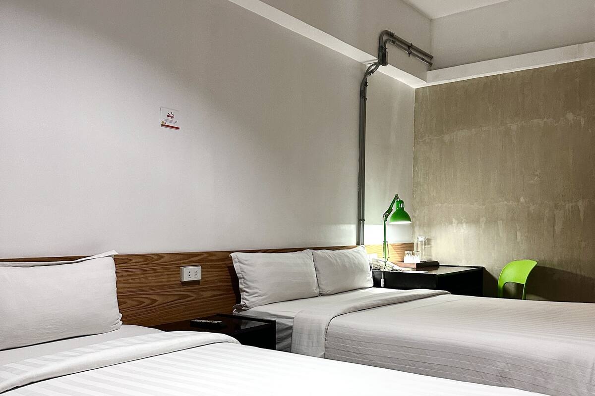A hotel room with two beds, a wooden headboard, a green chair, and a nightstand with a lamp.