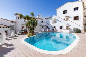 Une piscine entourée de bâtiments blancs aux toits de tuiles rouges, de palmiers et d’un ciel bleu azur.