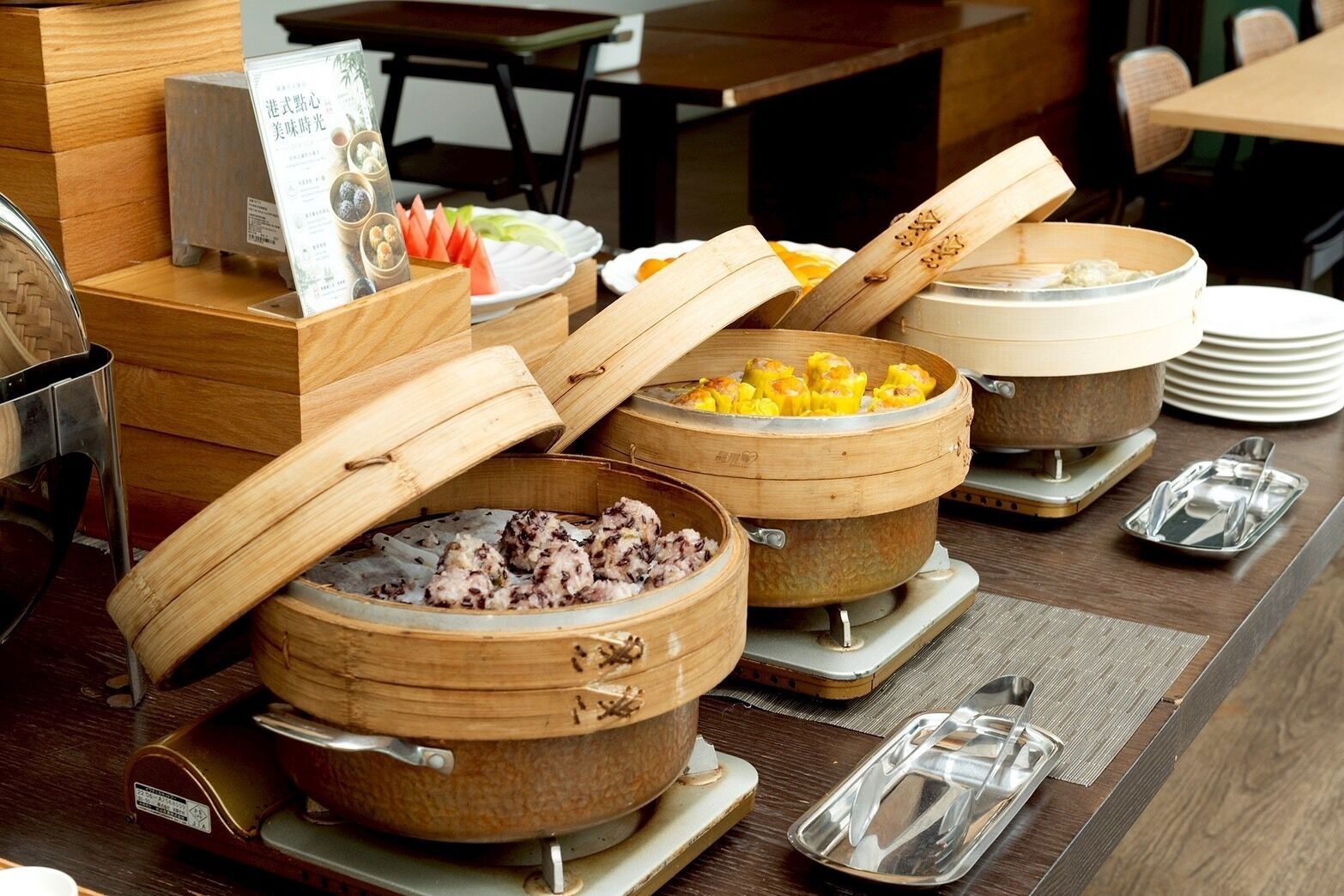 A buffet with various dishes in bamboo steamers, a sign with Japanese text, and a stack of plates.