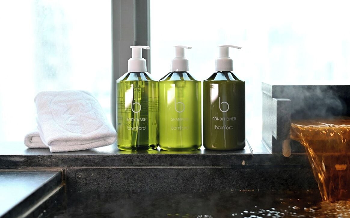 A bathroom with a towel, three bottles of body wash, shampoo, and conditioner, and a glass of amber liquid.