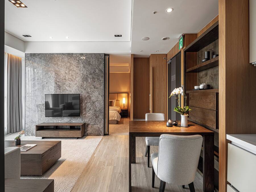 A modern living room with a marble wall, a flat-screen TV, a wooden dining table, and a kitchen area with wooden cabinets and a built-in oven.