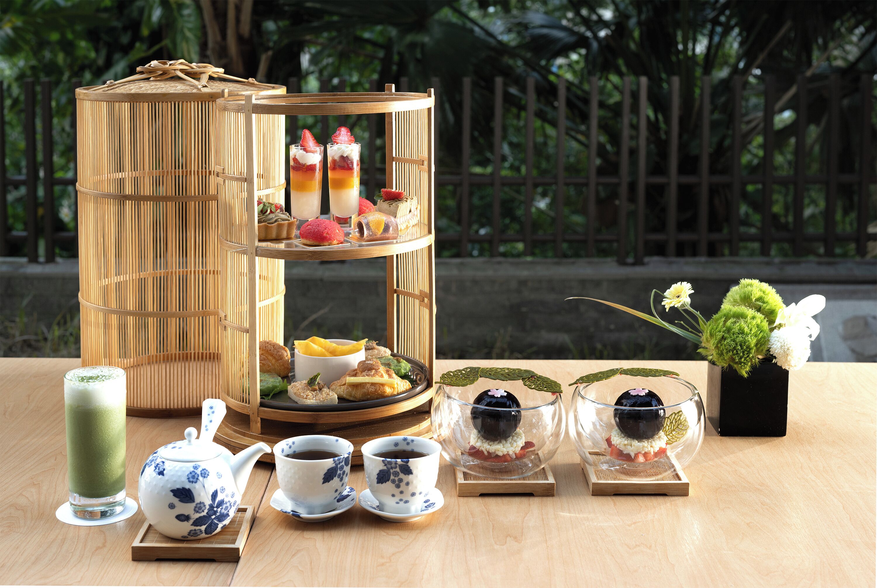 A bamboo tea set with a tiered stand holding desserts, a teapot, cups, and a glass of green tea.