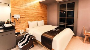 Ein Hotelzimmer mit einer Holzwand, einem Bett mit weißer Bettwäsche, einem schwarz-weißen Massagesessel, einem Schreibtisch mit Telefon und einem Fenster mit Blick auf den Nachthimmel.