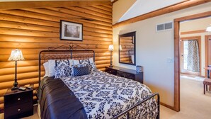 Un dormitorio con pared de troncos de madera, cama con ropa de cama estampada, lámparas de noche, un espejo y una puerta que da a otra habitación.