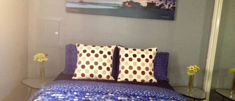A hotel room with a bed featuring a blue polka dot bedspread, two pillows with brown and white polka dots, a painting of a sunset on the wall, and two small tables with vases of yellow flowers.