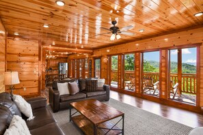 A spacious wooden cabin with a large living area featuring a sofa, a coffee table, and a dining table. There are large windows offering a view of the surrounding forest. - Lookout Lodge ~ Indoor Pool, Hot Tub & Mountain Views! (Sevierville)