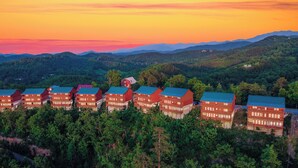 A row of red buildings with green roofs, surrounded by dense forest, set against a backdrop of mountains and a colorful sky. - Lookout Lodge ~ Indoor Pool, Hot Tub & Mountain Views! (Sevierville)