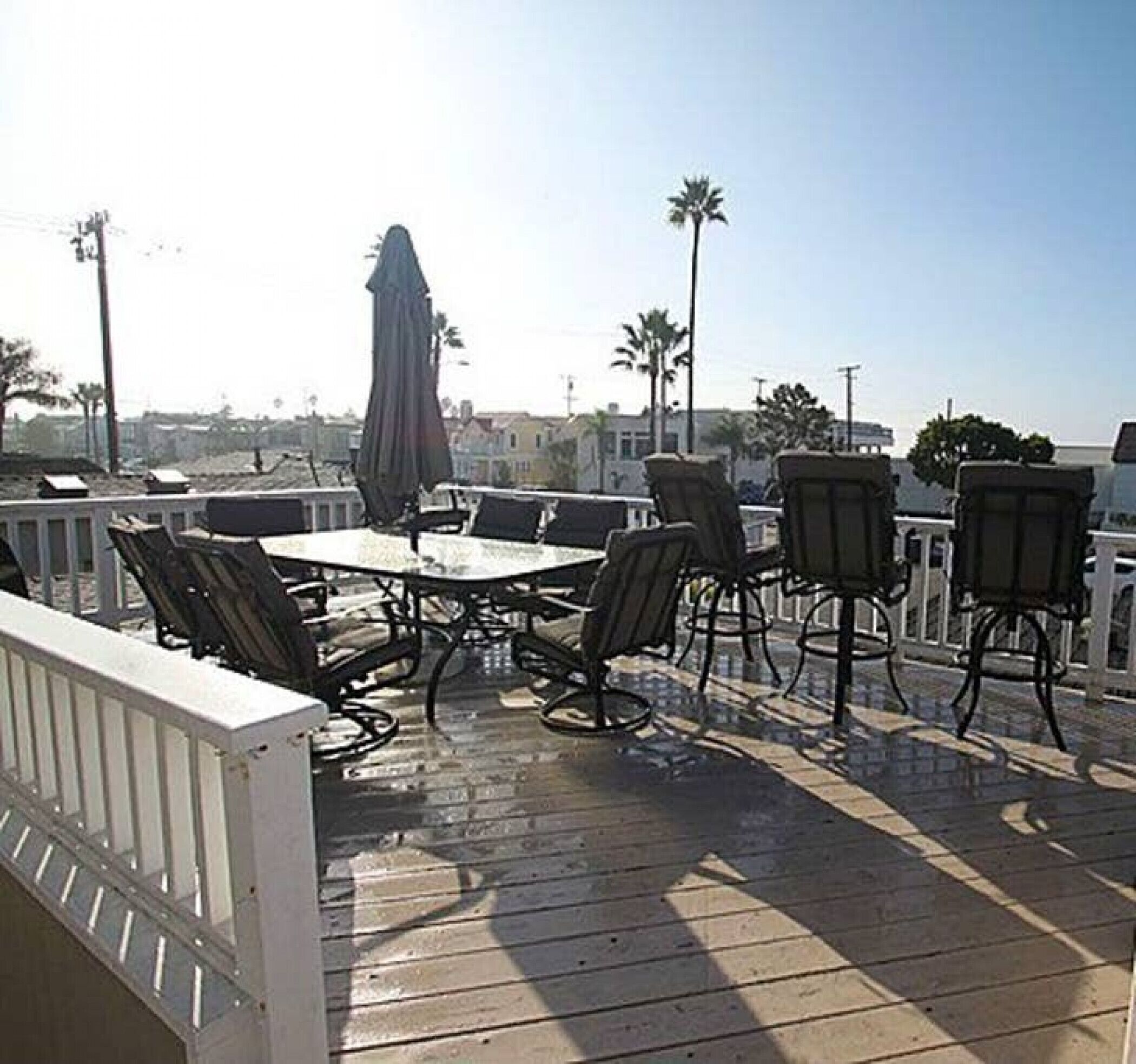 An outdoor patio with a table and chairs, a white railing, and palm trees in the background.
