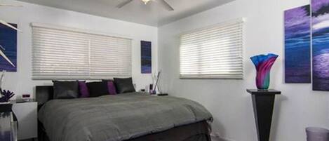 A bedroom with a bed, a ceiling fan, two windows with blinds, and a nightstand with a lamp.