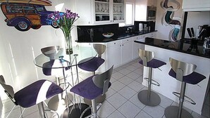 Une cuisine moderne avec des meubles blancs, un plan de travail noir et un plafond orné d’une fresque colorée. On y trouve un coin repas avec des chaises violettes et une table en verre.