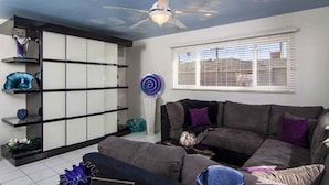 Un salon avec un ventilateur de plafond, un canapé gris et une fenêtre équipée de stores.
