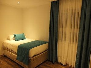 A single bed with a quilted headboard, a teal throw blanket, and a pillow. The room has wooden flooring and a window with sheer white curtains and a dark blue drape. - Cabañas en Puerto Varas (Puerto Varas)