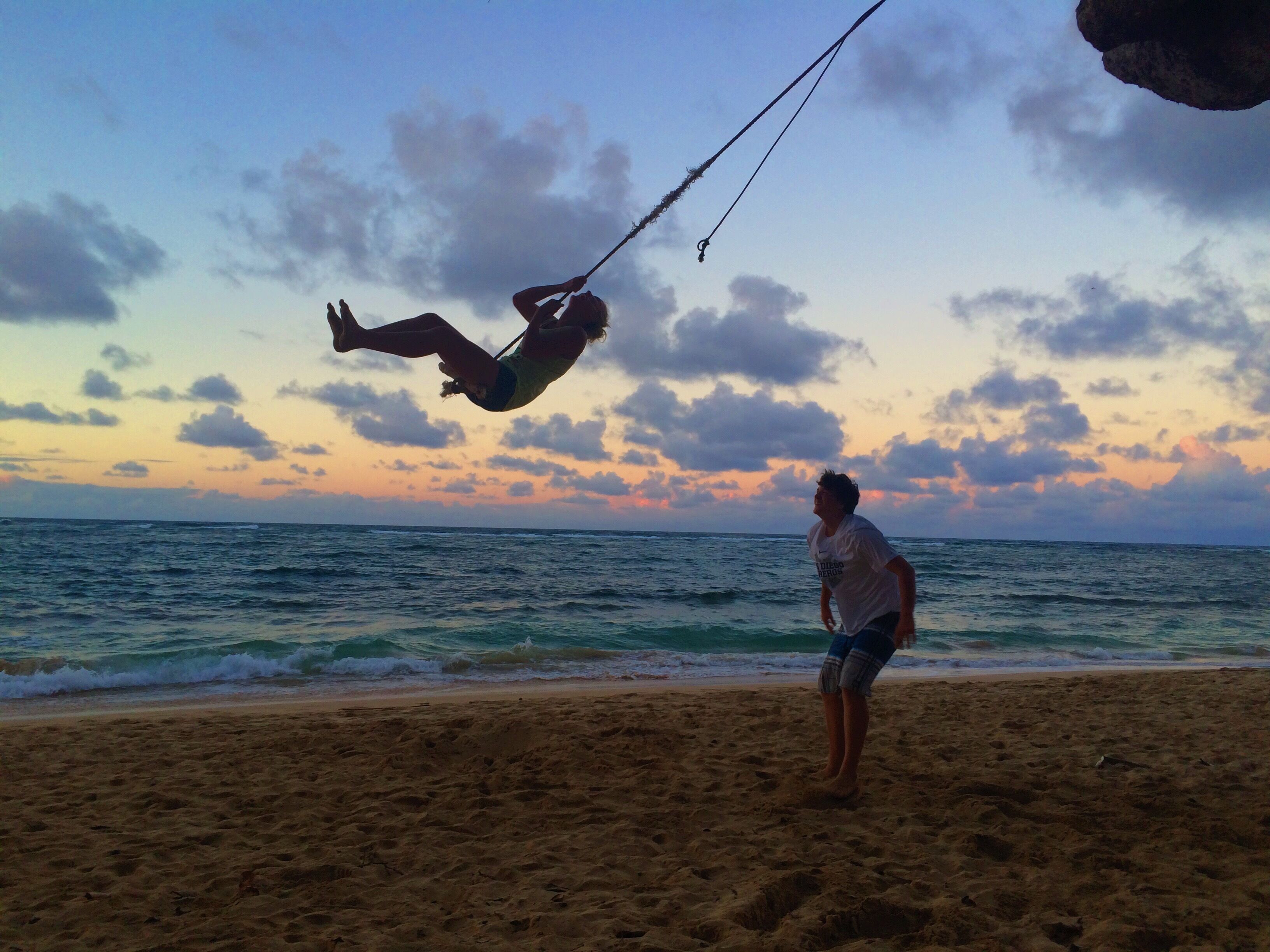 A beach scene with a person swinging on a rope over the water.