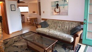 En stue med et sofabord i glas, en sofa med et mønster, et maleri af en strandscene, og et spiseområde med bord og stole.