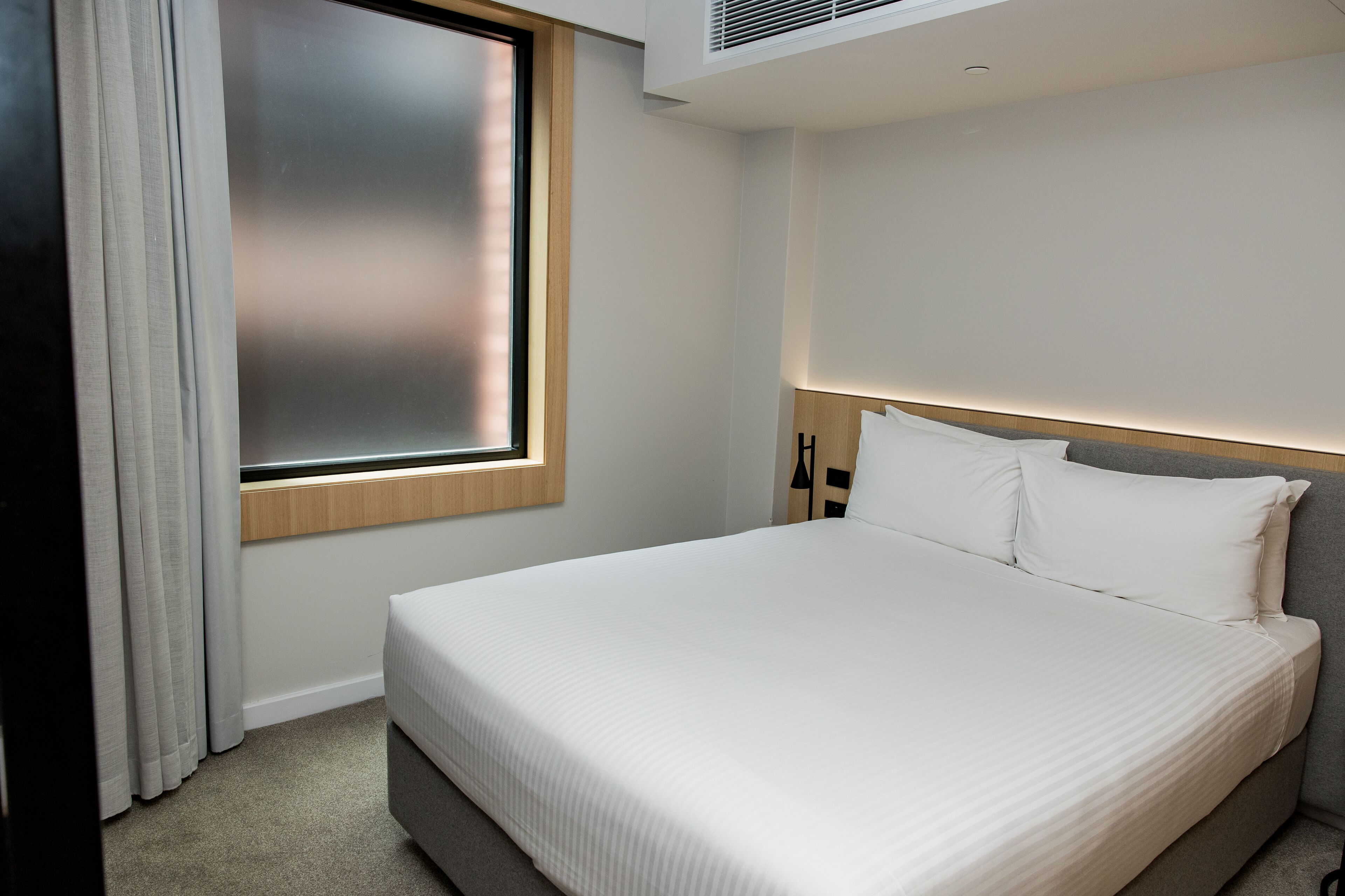 Una habitación de hotel moderna con una cama grande, una ventana con cortinas y un cabecero con iluminación integrada.